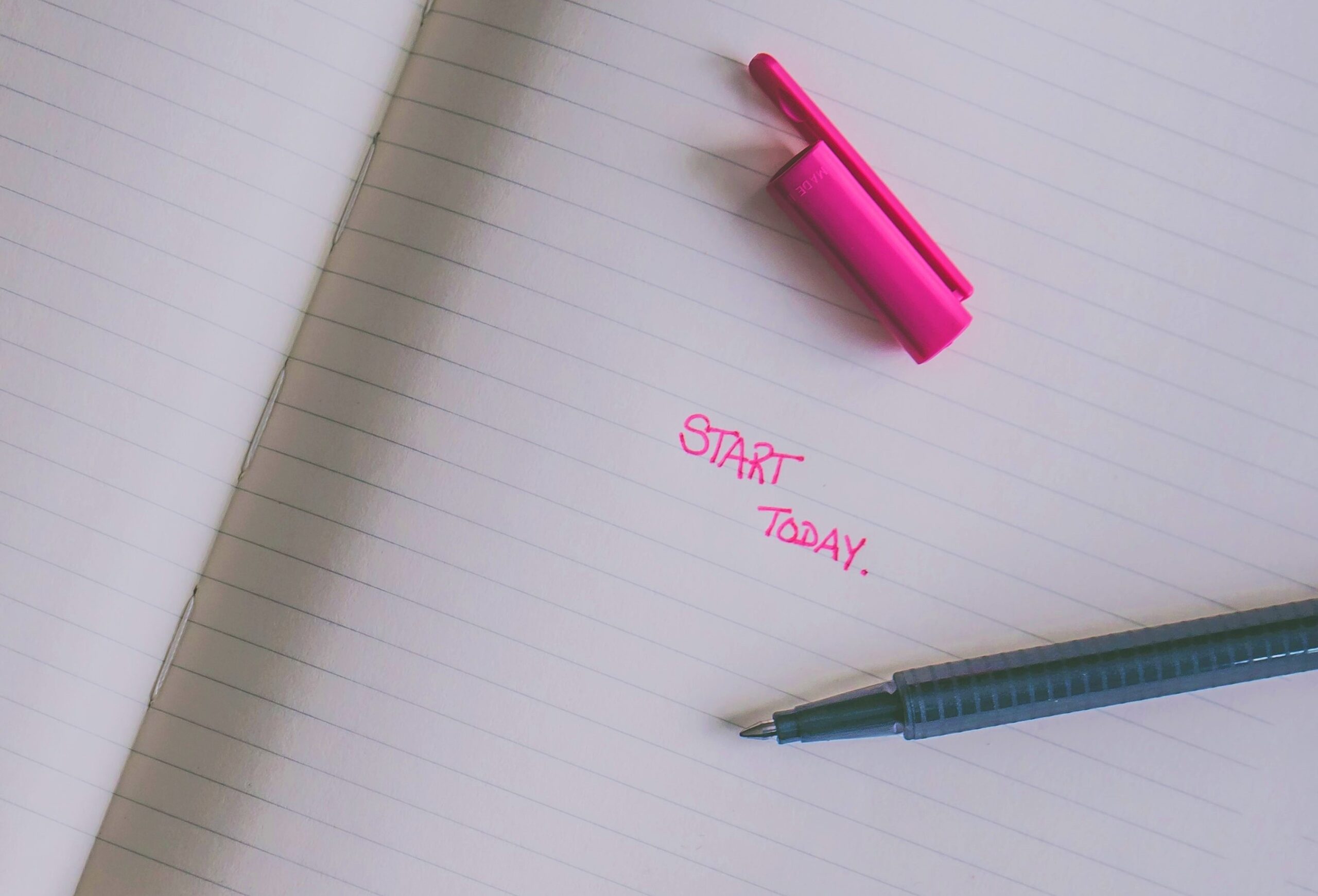 Image shows pink handwriting with words 'start today' on lined refill. A pen and pink pen cap lie next to the writing - people commonly try to create new habits with written intentions to help boost motivation and overcome procrastination. Photo-credits - by-Jessica-Lewis-on-Unsplash